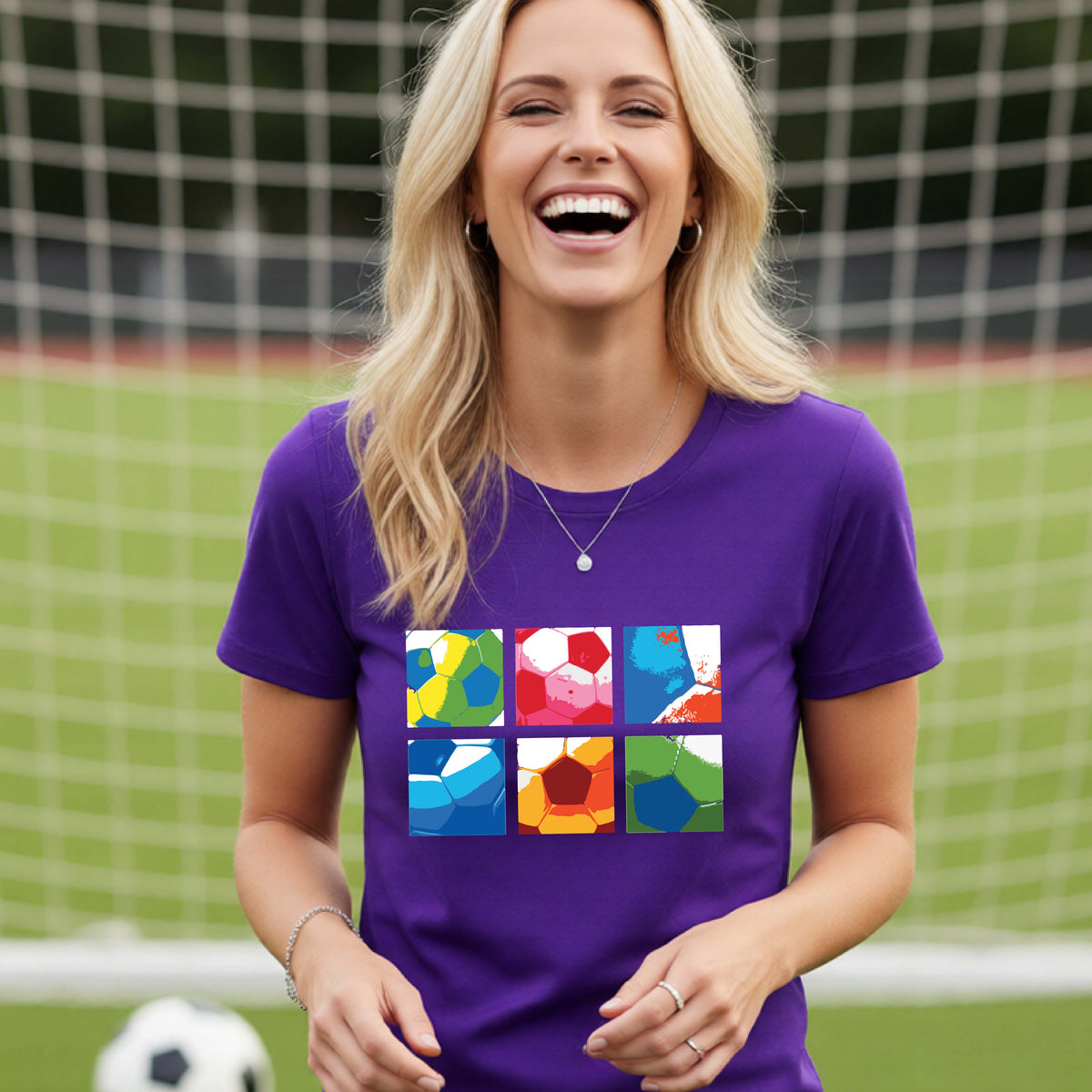 A smiling woman wearing a purple short-sleeve t-shirt standing on a soccer field in front of a goal net. with a colorful graphic of a 2x3 arrangement of squares of soccer balls, printed on the chest.
