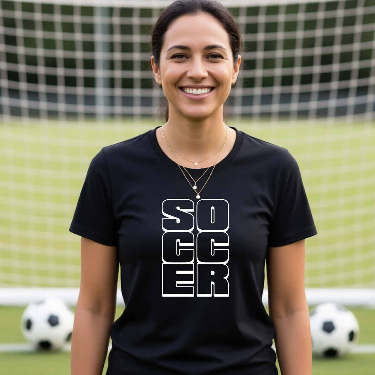 A smiling woman wearing a black short-sleeve t-shirt standing on a soccer field in front of a goal net. with a white graphic of the word ‘SOCCER’ stacked in a 2x3 arrangement, printed on the chest.