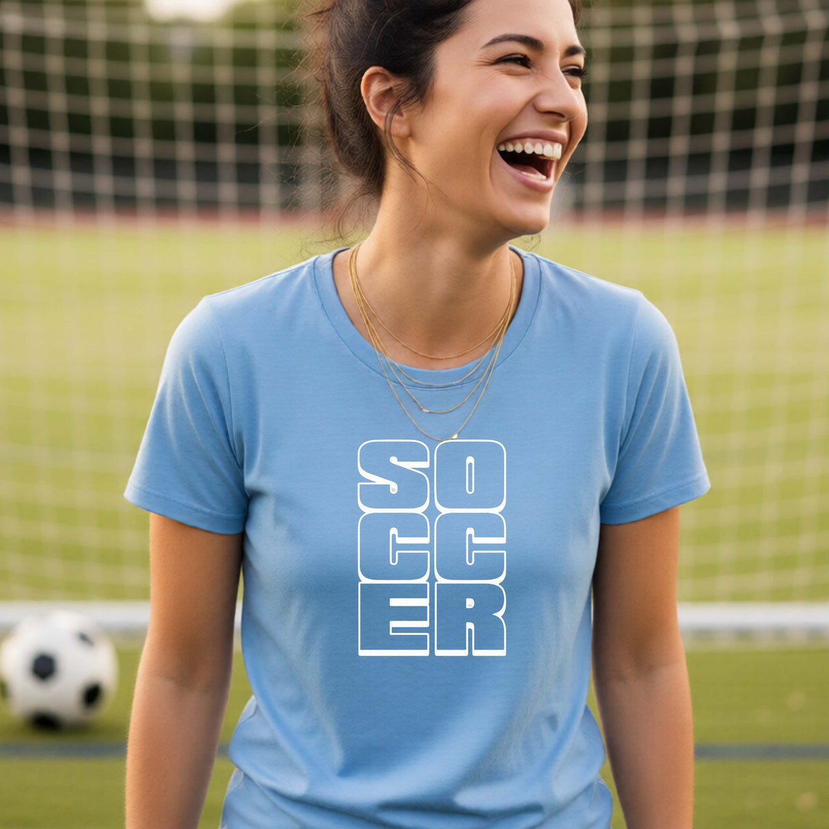 A smiling woman wearing a blue short-sleeve t-shirt standing on a soccer field in front of a goal net. with a white graphic of the word ‘SOCCER’ stacked in a 2x3 arrangement, printed on the chest.