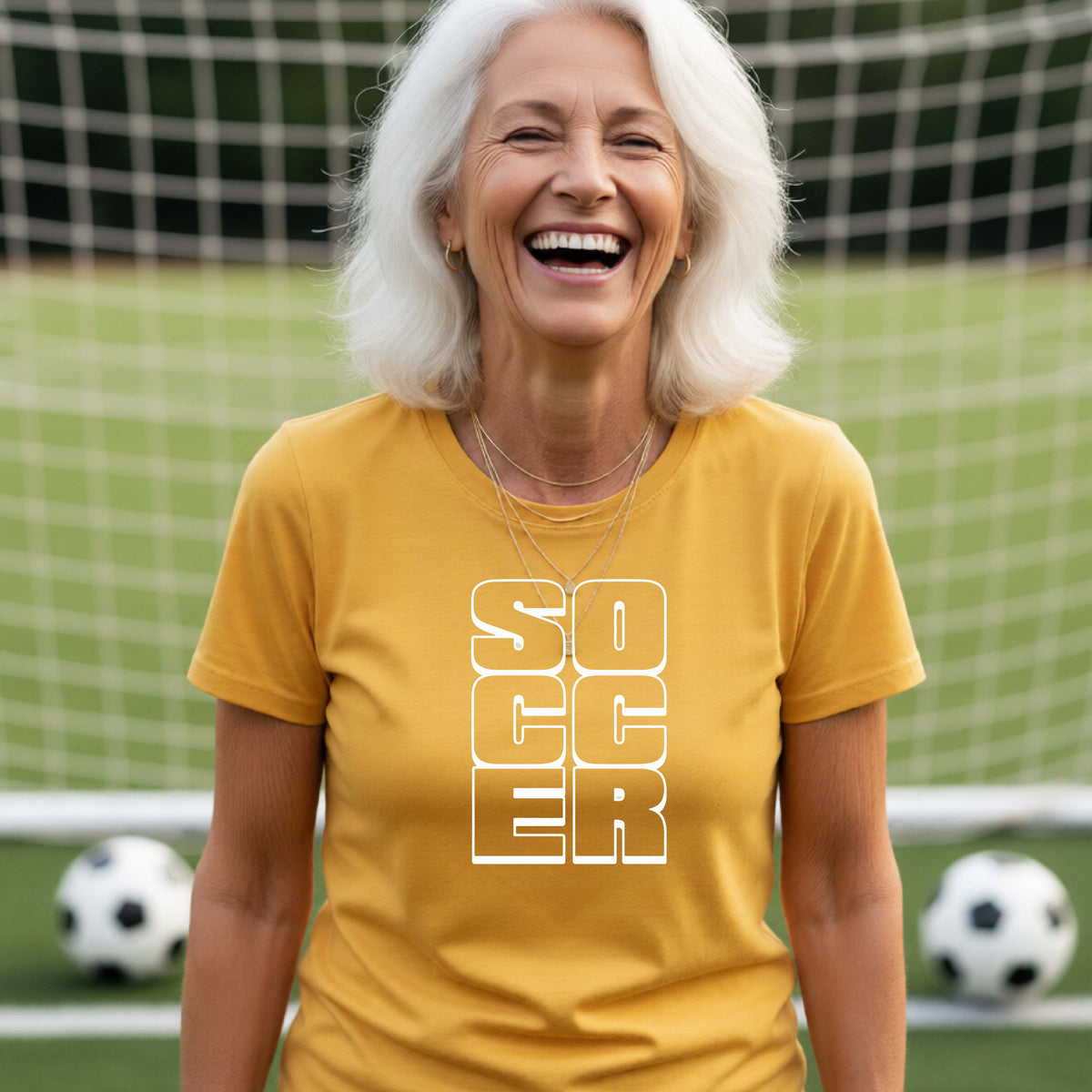 A smiling woman wearing a gold short-sleeve t-shirt standing on a soccer field in front of a goal net. with a white graphic of the word ‘SOCCER’ stacked in a 2x3 arrangement, printed on the chest.