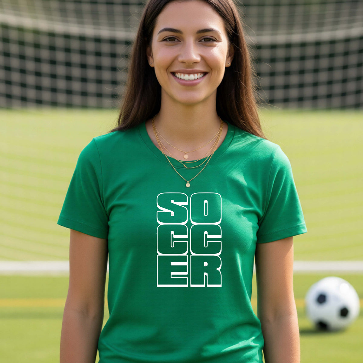 A smiling woman wearing a green short-sleeve t-shirt standing on a soccer field in front of a goal net. with a white graphic of the word ‘SOCCER’ stacked in a 2x3 arrangement, printed on the chest.