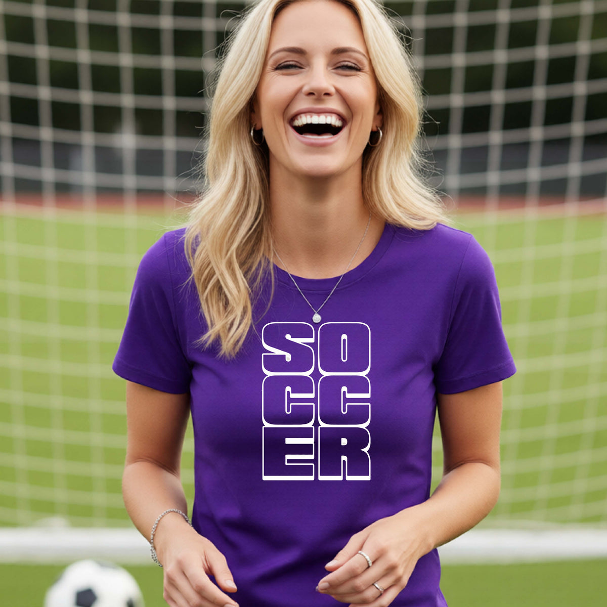 A smiling woman wearing a purple short-sleeve t-shirt standing on a soccer field in front of a goal net. with a white graphic of the word ‘SOCCER’ stacked in a 2x3 arrangement, printed on the chest.