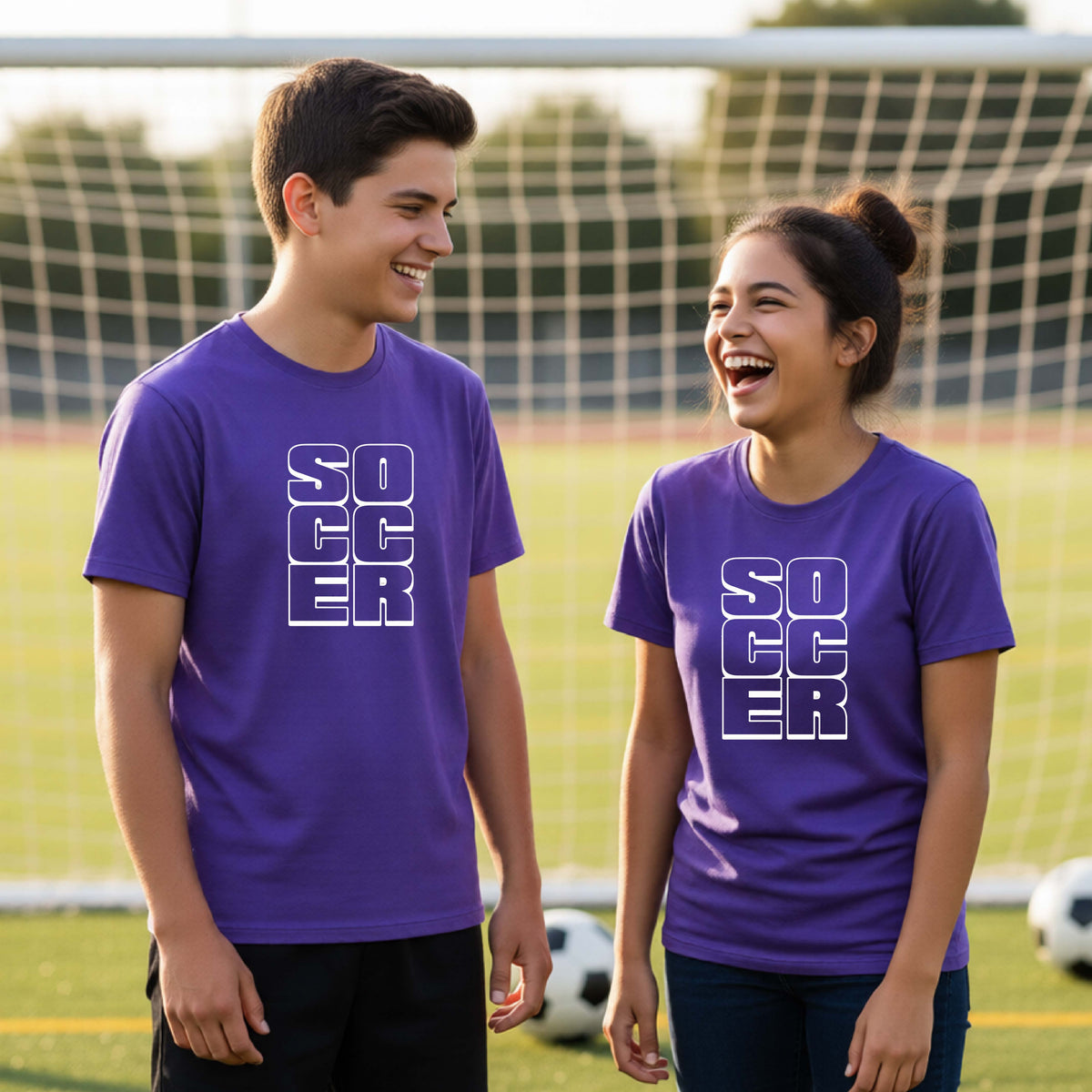 Two young athletes wearing matching purple t-shirts, standing in front of a soccr goal and balls, with a white graphic of the word ‘SOCCER’ stacked in a 2x3 arrangement, printed on the chest.