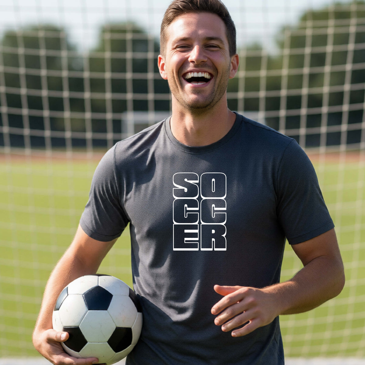 A smiling man wearing a dark gray short-sleeve t-shirt standing on a soccer field in front of a goal net. with a white graphic of the word ‘SOCCER’ stacked in a 2x3 arrangement, printed on the chest.