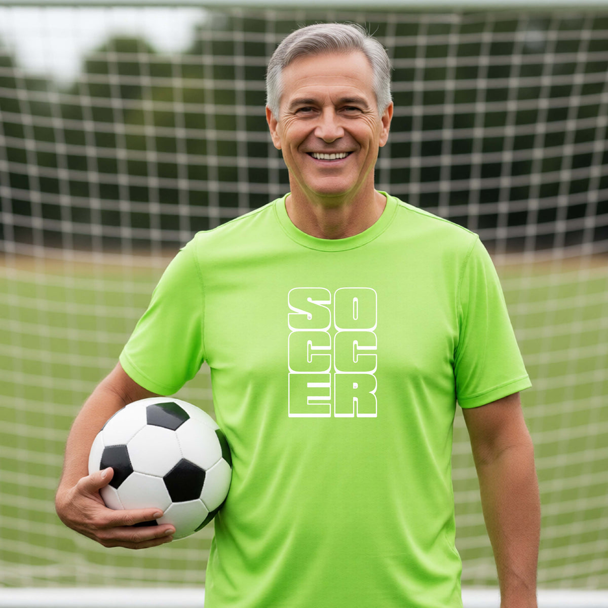 A smiling man wearing a lime green short-sleeve t-shirt standing on a soccer field in front of a goal net. with a white graphic of the word ‘SOCCER’ stacked in a 2x3 arrangement, printed on the chest.