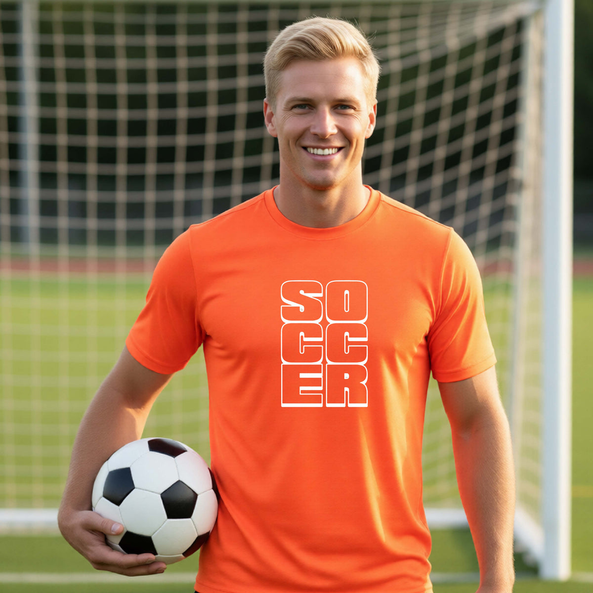 A smiling man wearing an orange short-sleeve t-shirt standing on a soccer field in front of a goal net. with a white graphic of the word ‘SOCCER’ stacked in a 2x3 arrangement, printed on the chest.