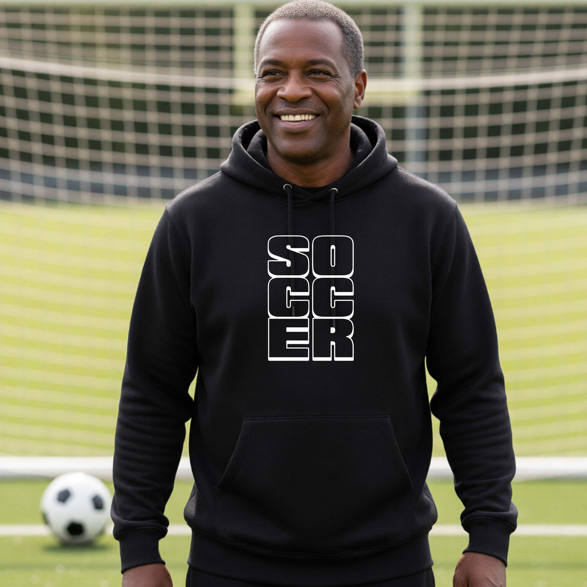 A smiling man wearing a black hoodie pullover standing on a soccer field in front of a goal net. with a white graphic of the word ‘SOCCER’ stacked in a 2x3 arrangement, printed on the chest.
