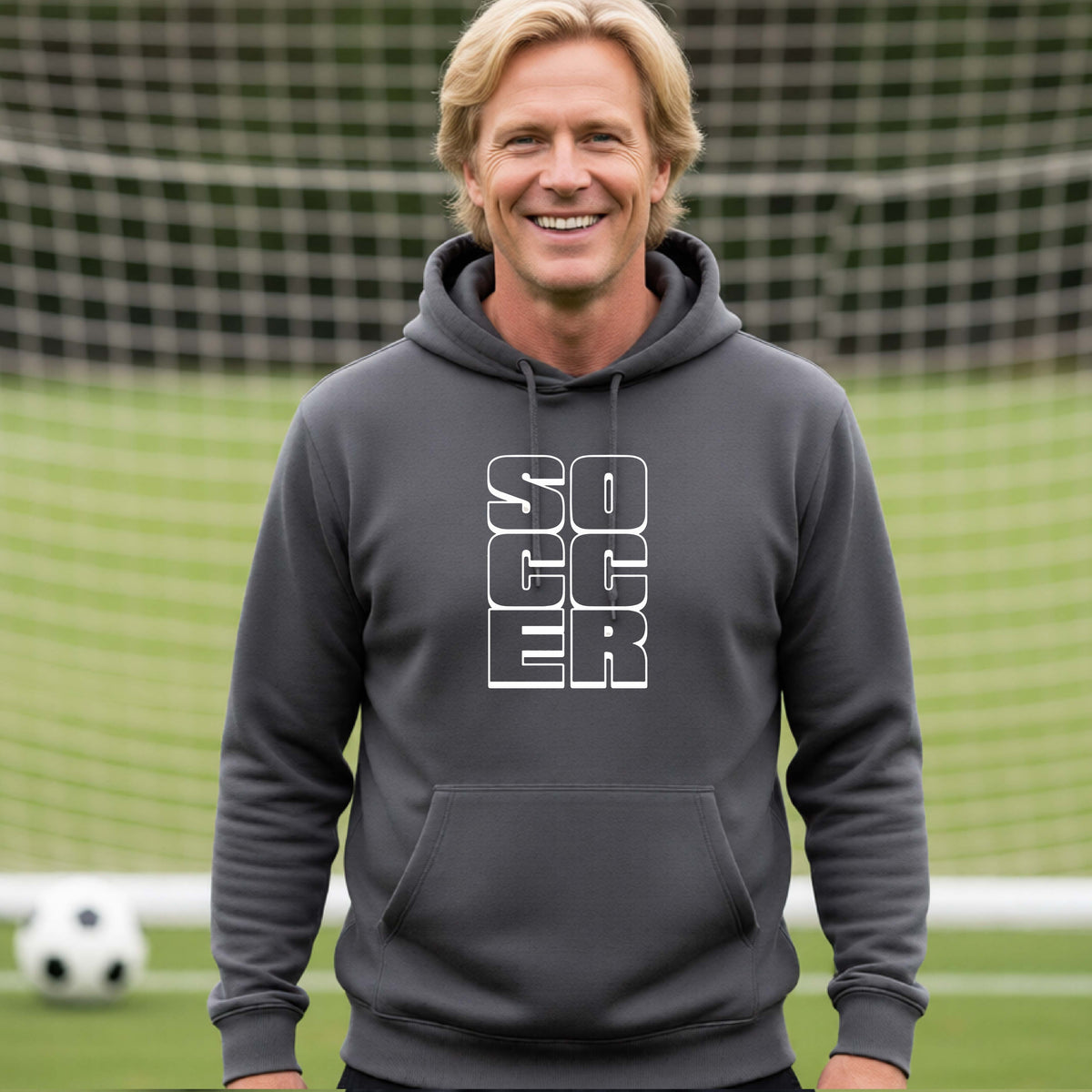 A smiling man wearing a gray hoodie pullover standing on a soccer field in front of a goal net. with a white graphic of the word ‘SOCCER’ stacked in a 2x3 arrangement, printed on the chest.