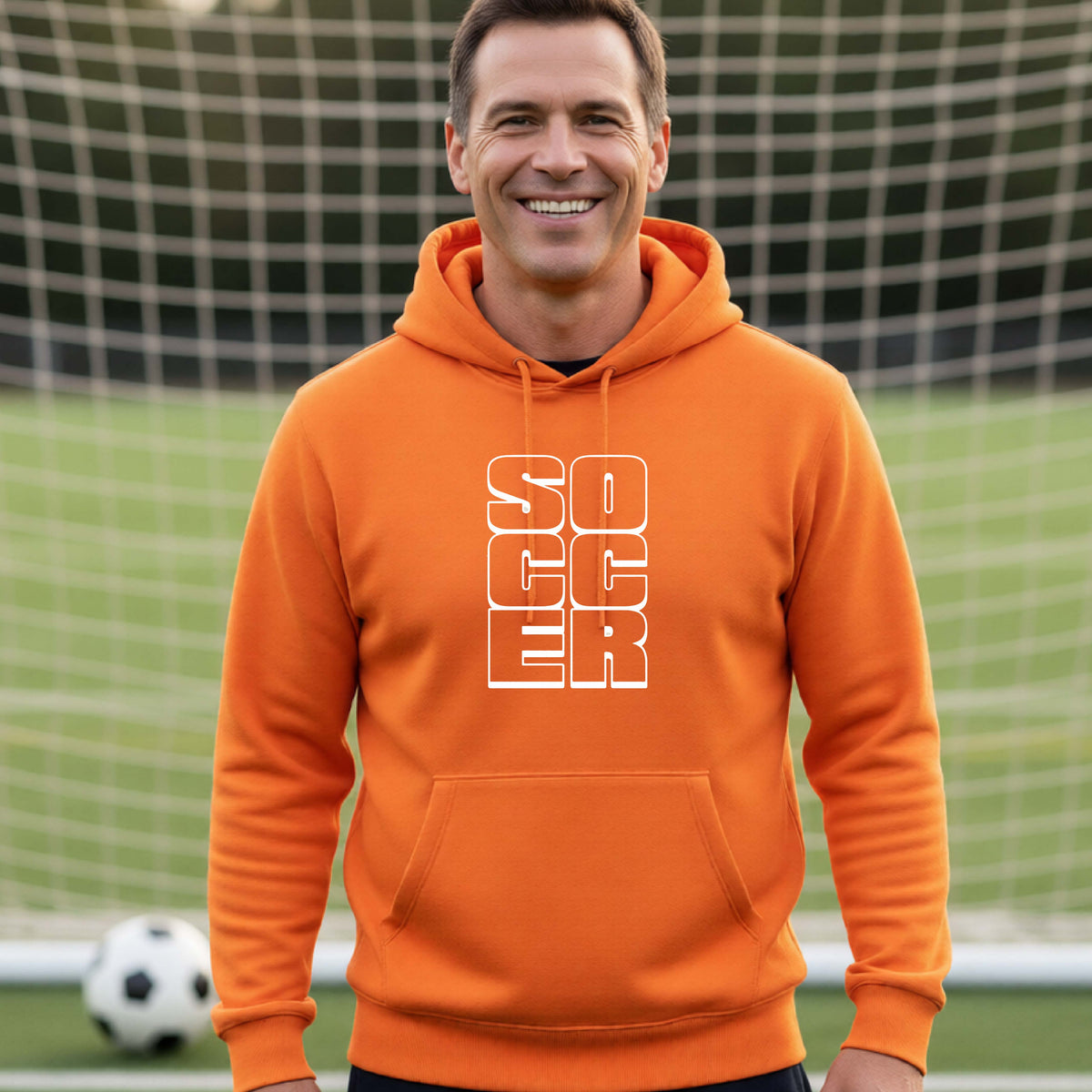 A smiling man wearing an orange hoodie pullover standing on a soccer field in front of a goal net. with a white graphic of the word ‘SOCCER’ stacked in a 2x3 arrangement, printed on the chest.
