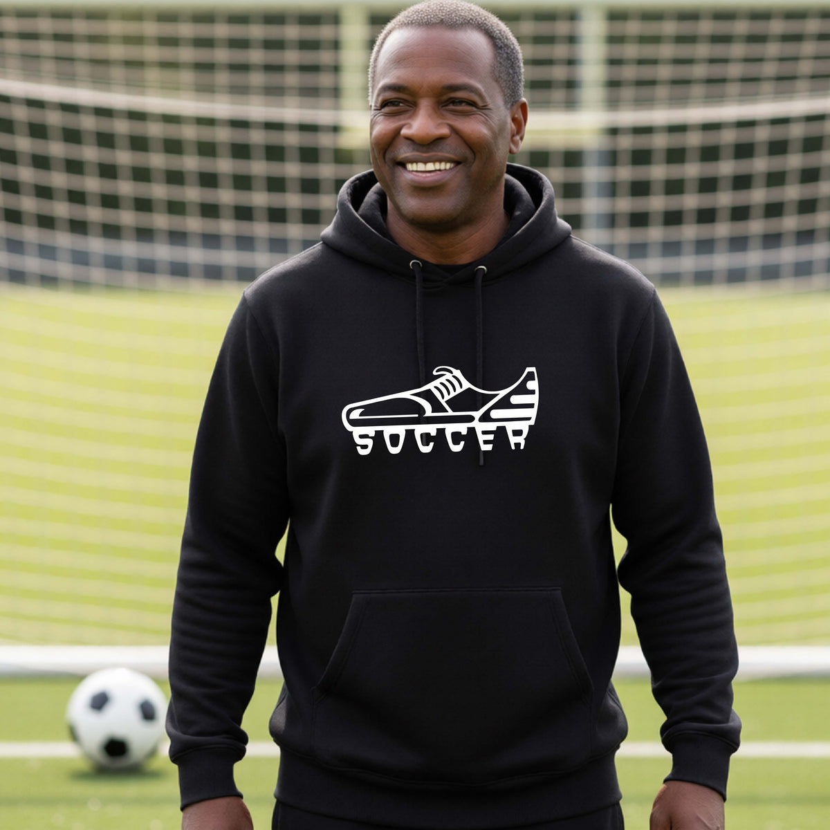 A smiling man, standing on a soccer field in front of a goal net. wearing a black hoodie pullover with a white soccer cleat graphic and the word 'SOCCER' printed on the chest.