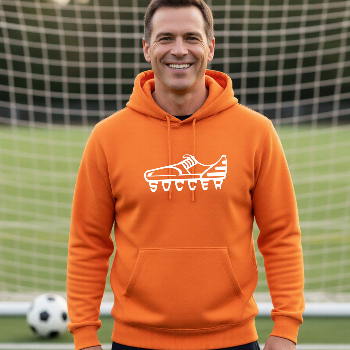 A smiling man, standing on a soccer field in front of a goal net. wearing an orange hoodie pullover with a white soccer cleat graphic and the word 'SOCCER' printed on the chest.