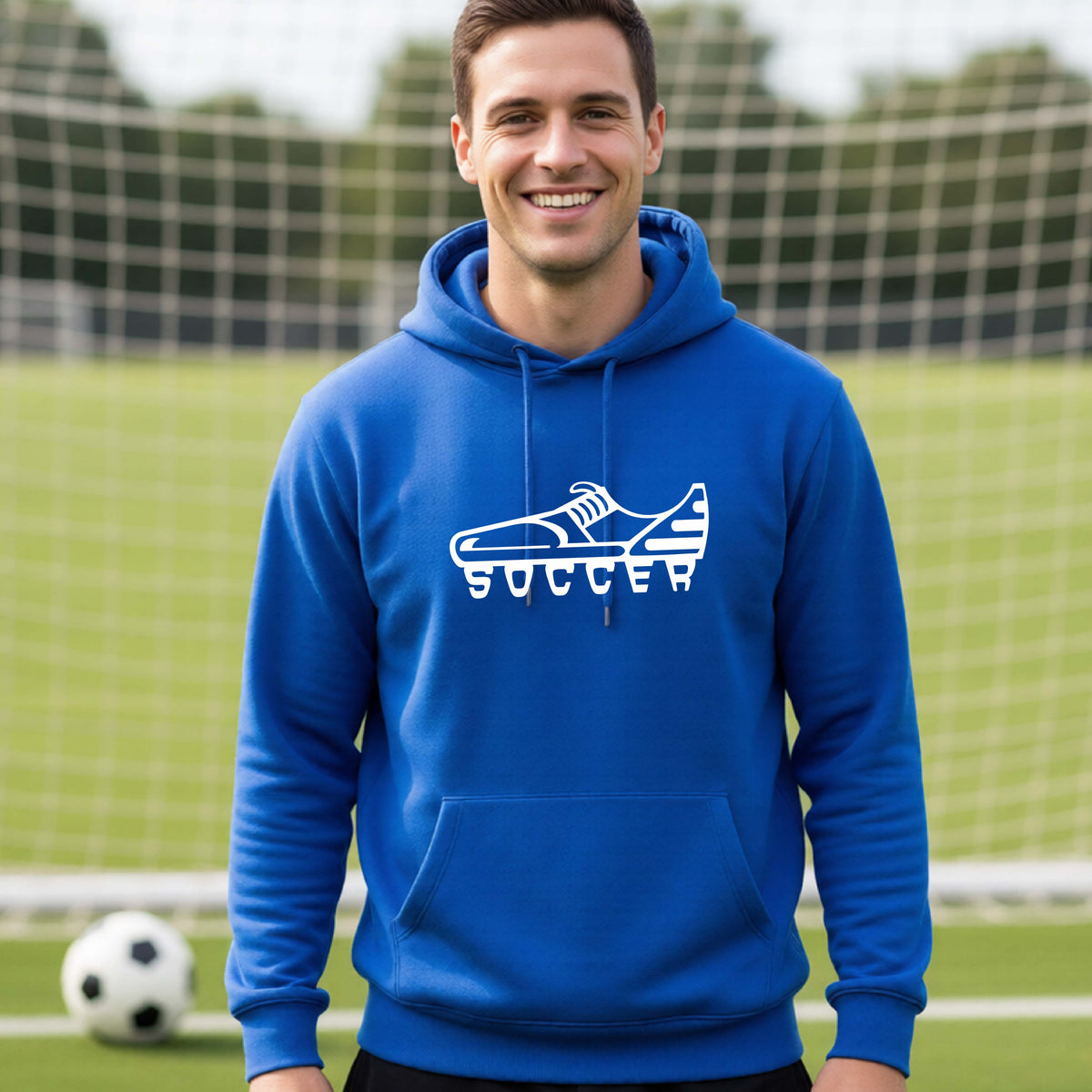 A smiling man, standing on a soccer field in front of a goal net. wearing a royal blue hoodie pullover with a white soccer cleat graphic and the word 'SOCCER' printed on the chest.