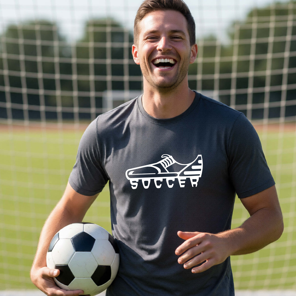 A smiling man on a soccer field wearing a dark gray t-shirt with a white soccer cleat graphic and the word 'SOCCER' underneath, holding a black and white soccer ball in front of a soccer goal.