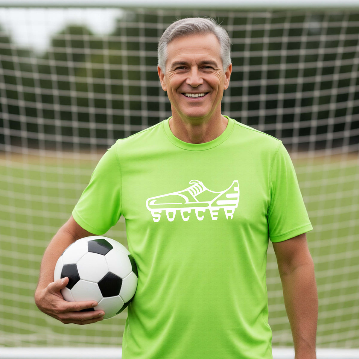 A smiling man on a soccer field wearing a lime green t-shirt with a white soccer cleat graphic and the word 'SOCCER' underneath, holding a black and white soccer ball in front of a soccer goal.