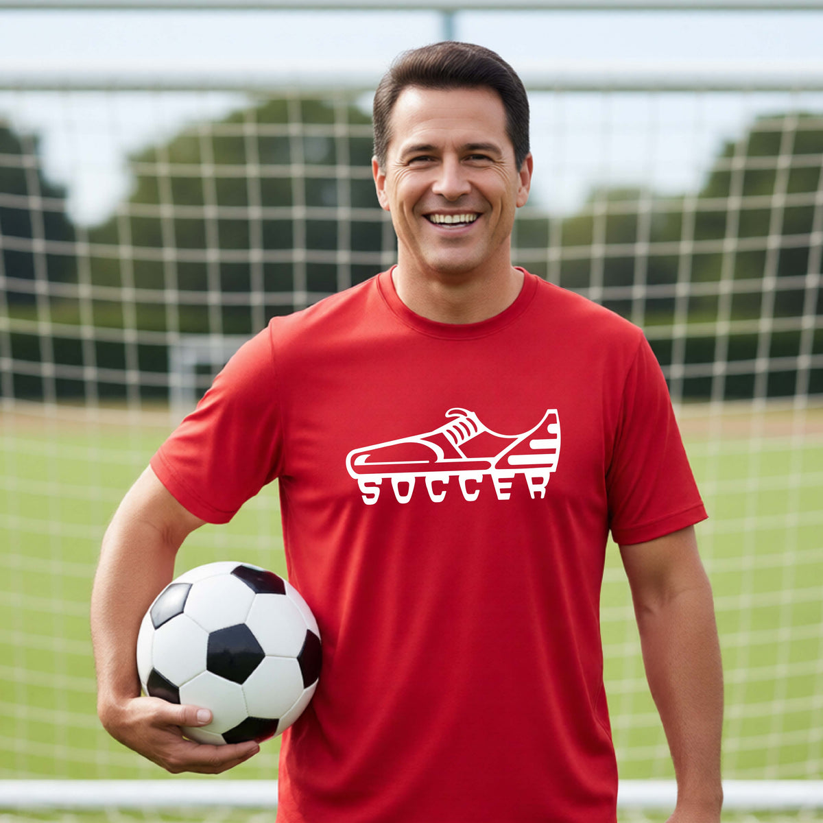 A smiling man on a soccer field wearing a red t-shirt with a white soccer cleat graphic and the word 'SOCCER' underneath, holding a black and white soccer ball in front of a soccer goal.