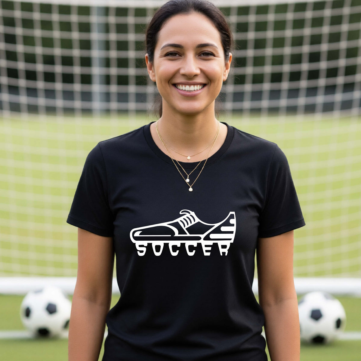A smiling woman, standing on a soccer field in front of a goal net. wearing a black t-shirt with a white soccer cleat graphic and the word 'SOCCER' printed on the chest.