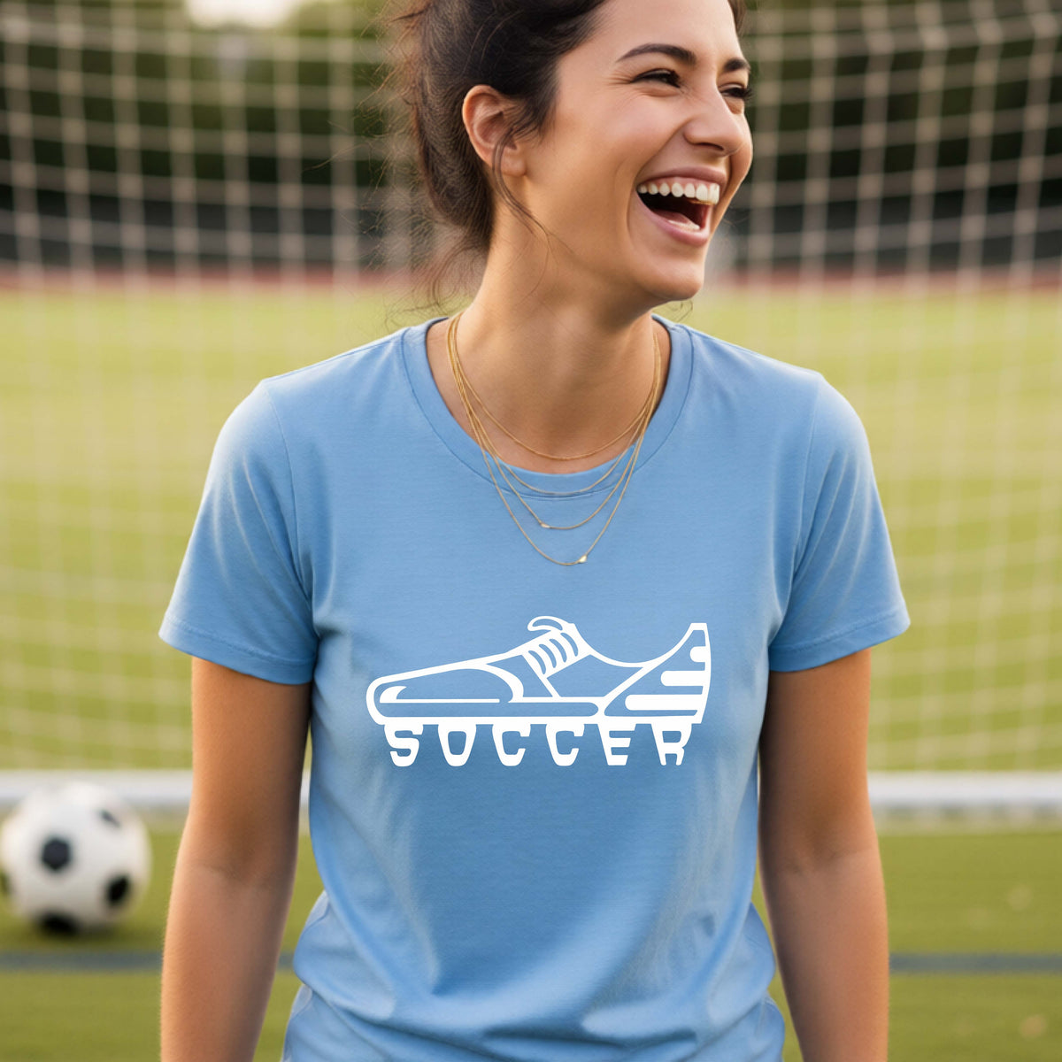 A smiling woman, standing on a soccer field in front of a goal net. wearing a blue t-shirt with a white soccer cleat graphic and the word 'SOCCER' printed on the chest.