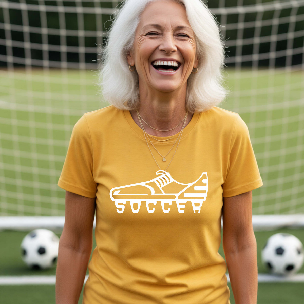 A smiling woman, standing on a soccer field in front of a goal net. wearing a gold t-shirt with a white soccer cleat graphic and the word 'SOCCER' printed on the chest.