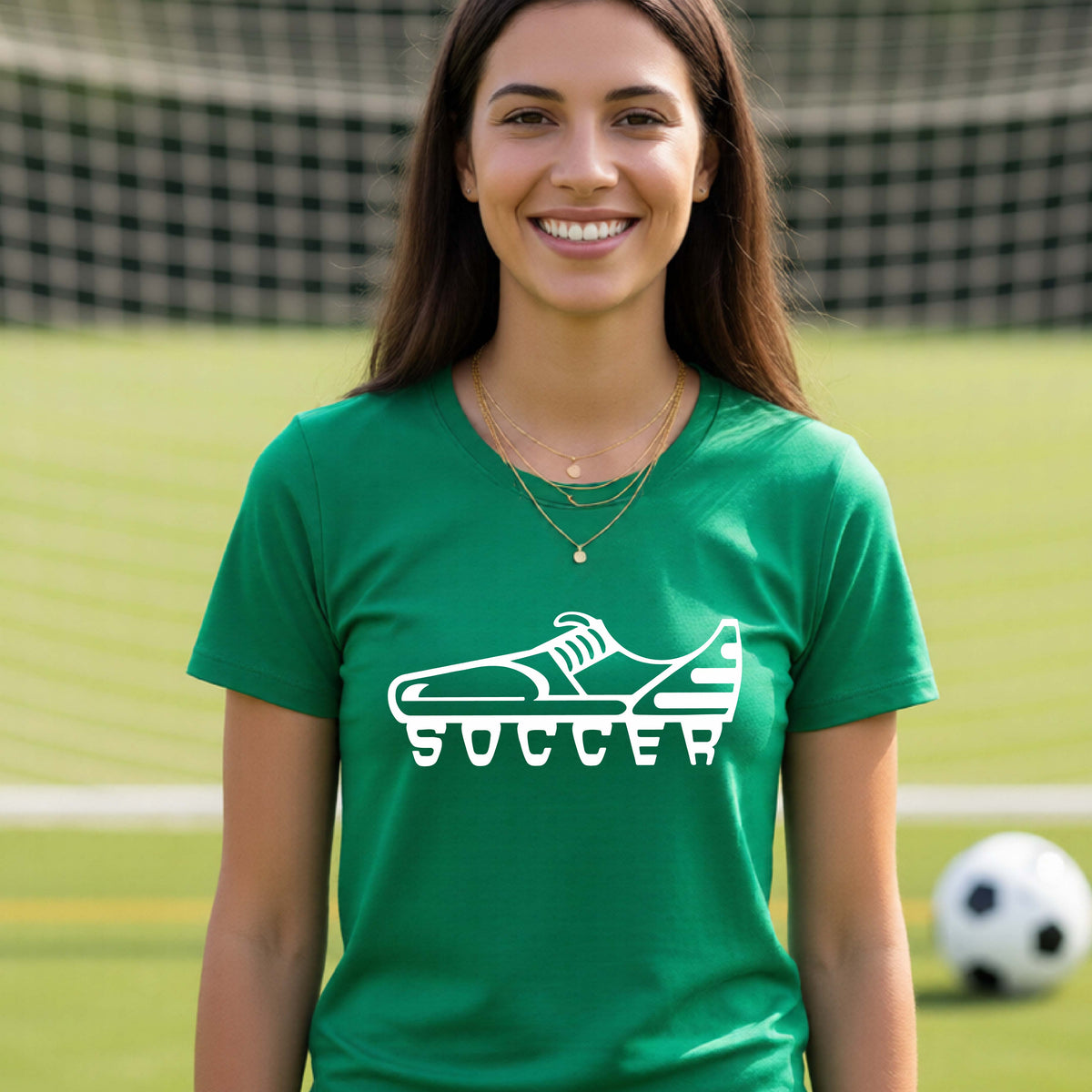 A smiling woman, standing on a soccer field in front of a goal net. wearing a green t-shirt with a white soccer cleat graphic and the word 'SOCCER' printed on the chest.