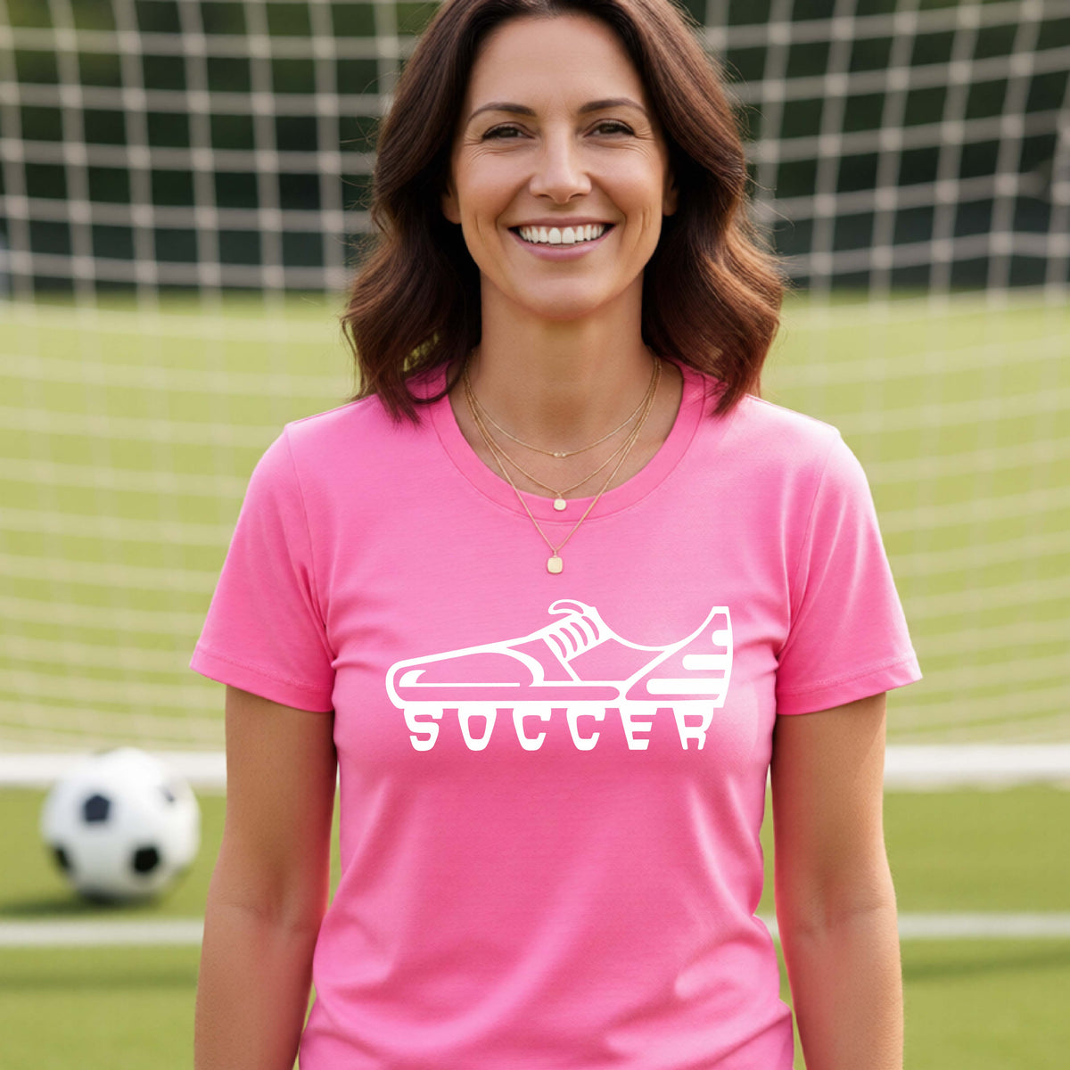 A smiling woman, standing on a soccer field in front of a goal net. wearing a pink t-shirt with a white soccer cleat graphic and the word 'SOCCER' printed on the chest.