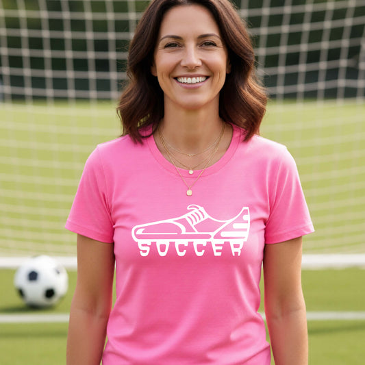 A smiling woman, standing on a soccer field in front of a goal net. wearing a pink t-shirt with a white soccer cleat graphic and the word 'SOCCER' printed on the chest.