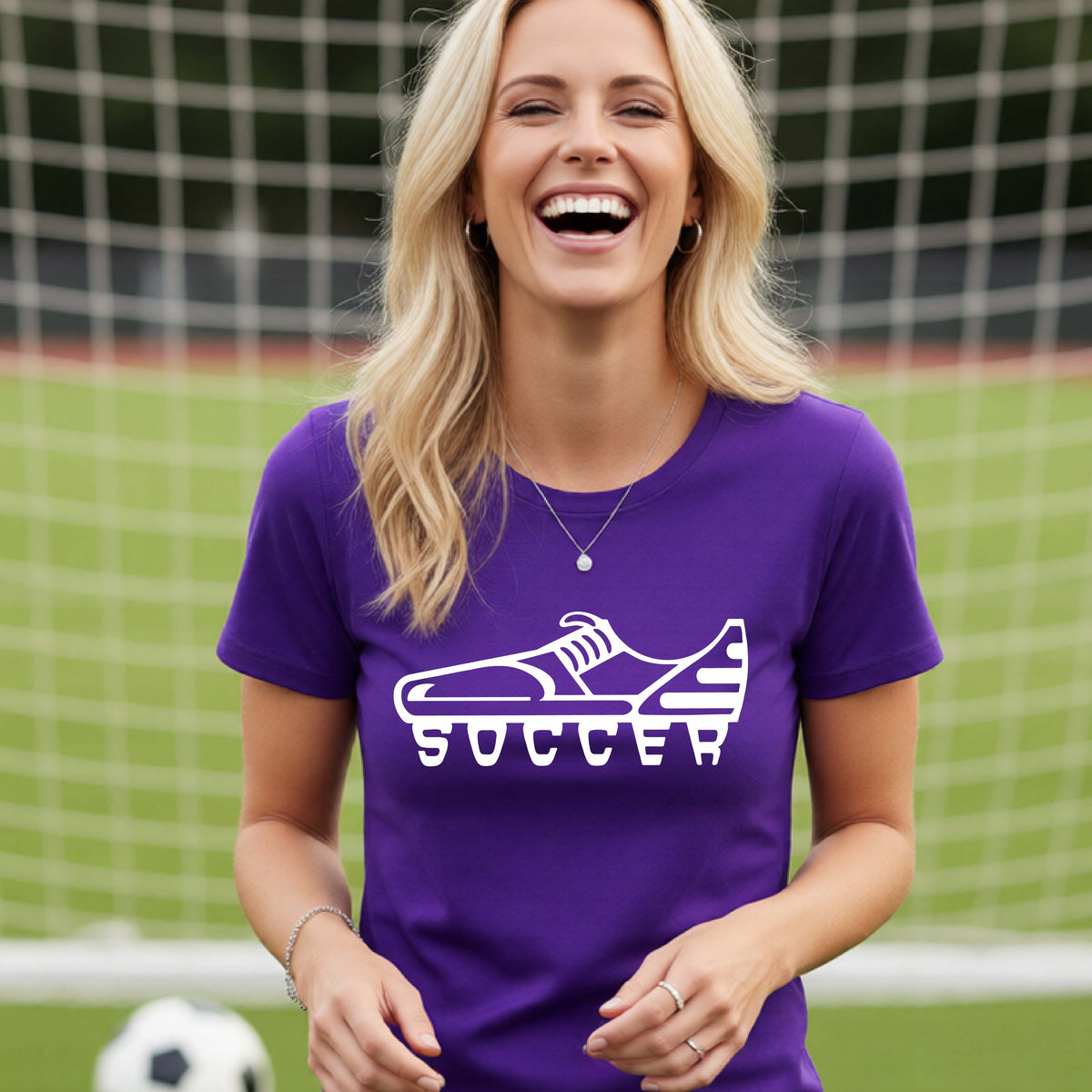 A smiling woman, standing on a soccer field in front of a goal net. wearing a purple t-shirt with a white soccer cleat graphic and the word 'SOCCER' printed on the chest.