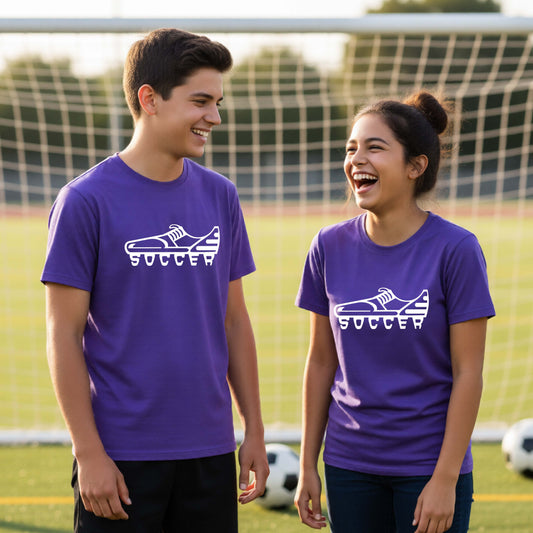 Two smiling young athletes, a boy and a girl, standing on a soccer field in front of a goal net. Both are wearing matching purple t-shirts with a white soccer cleat graphic and the word 'SOCCER' printed on the chest.