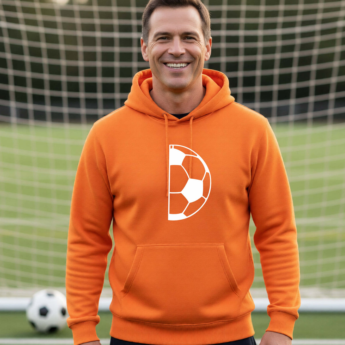 A smiling man wearing an orange hoodie with a white soccer ball graphic on the chest, standing on a soccer field in front of a goal net.