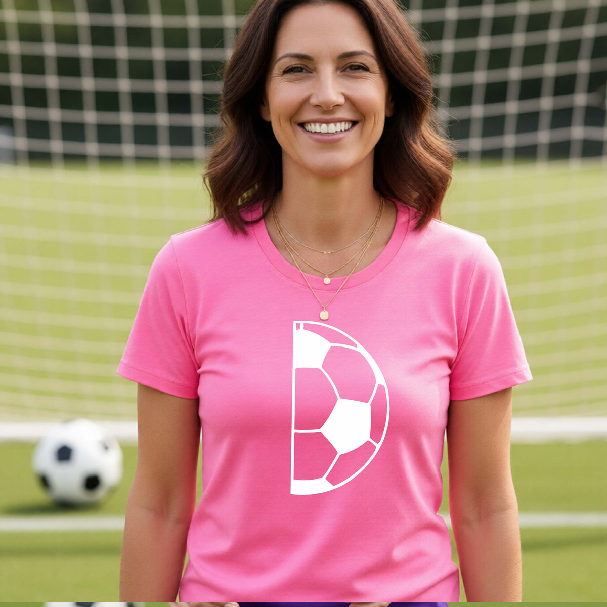 A woman laughing on a soccer field wearing a pink t-shirt with a white half soccer ball graphic inside a semicircle on the chest.
