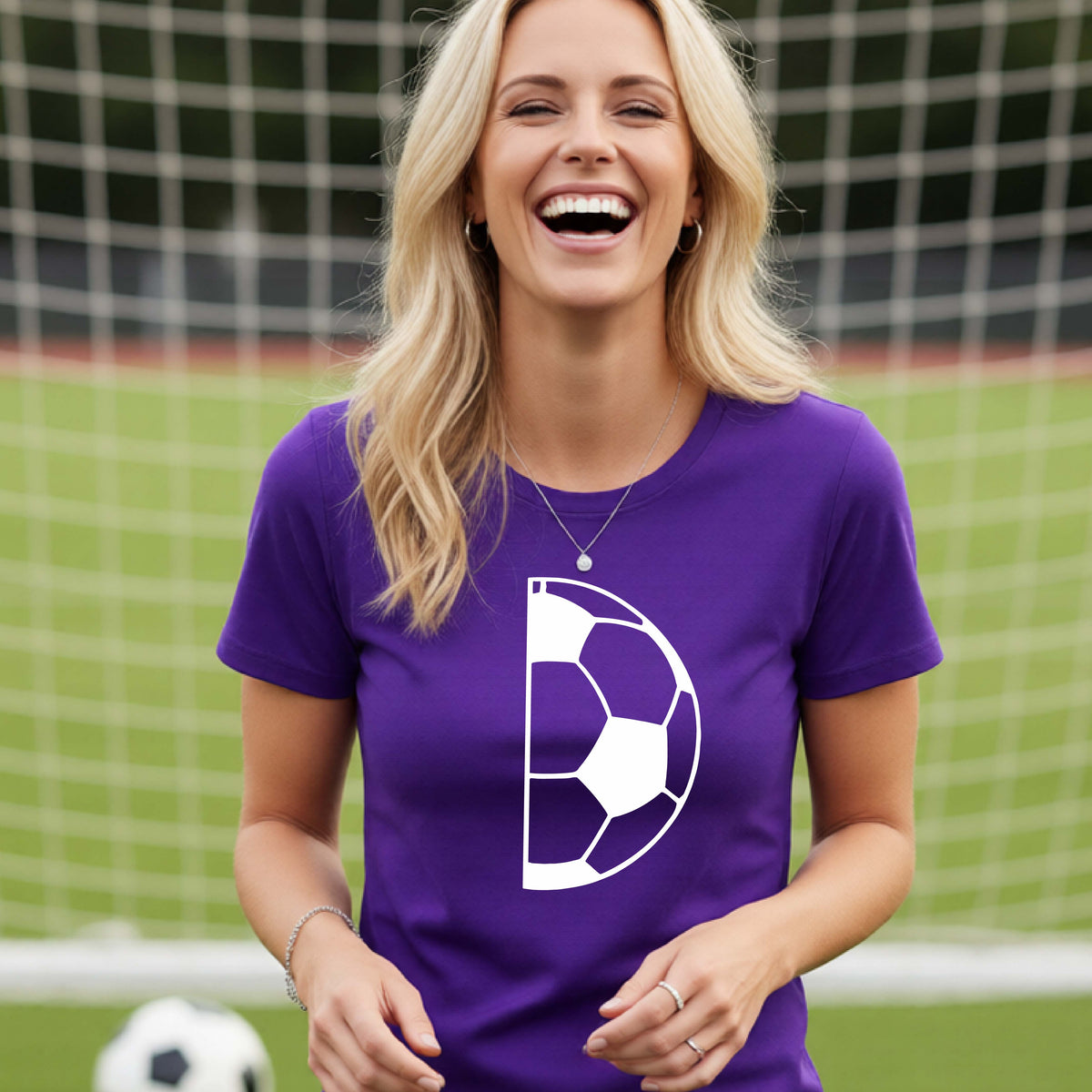 A woman laughing on a soccer field wearing a purple t-shirt with a white half soccer ball graphic inside a semicircle on the chest.