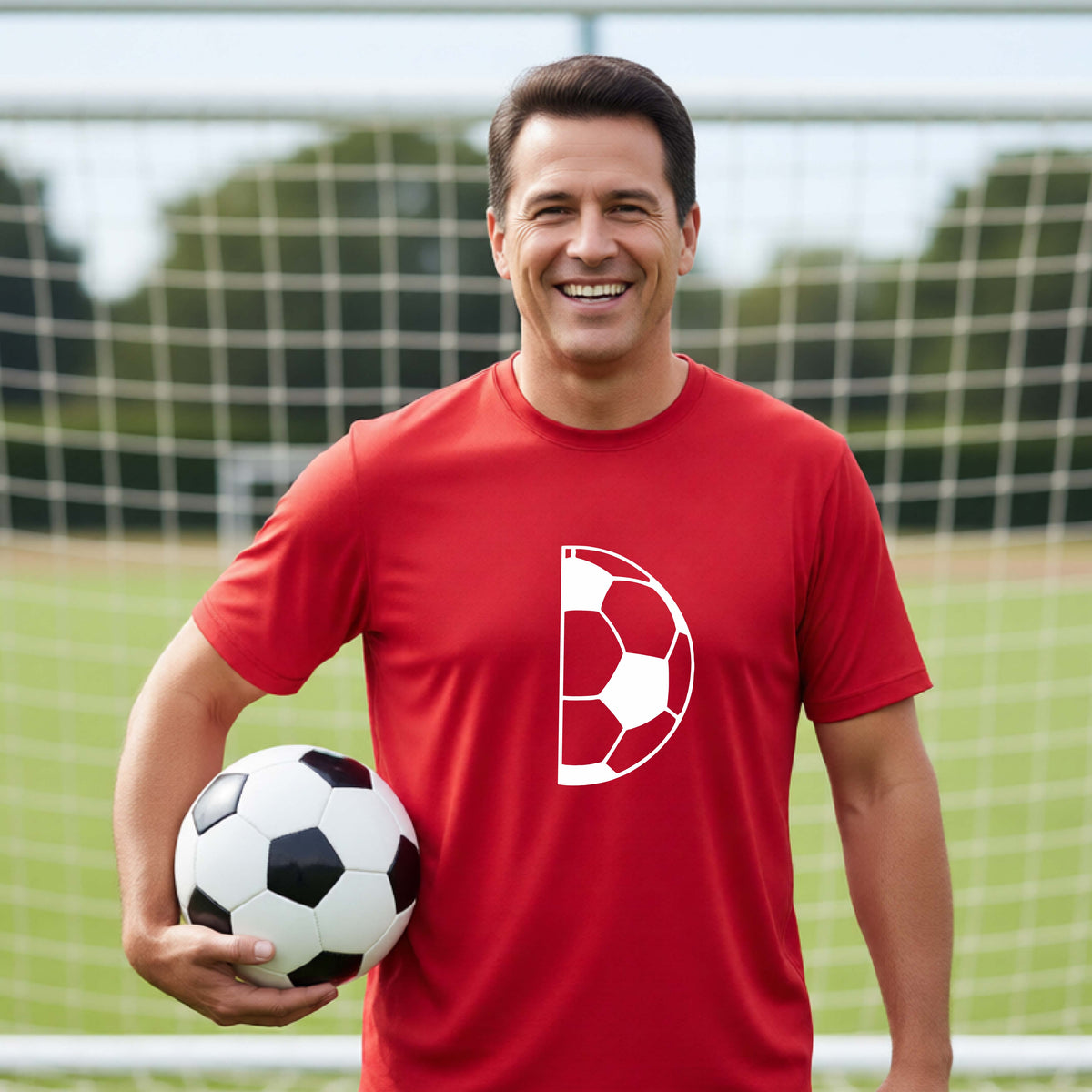 A smiling older man wearing a red short-sleeve t-shirt with a white half soccer ball graphic, standing on a green soccer field in front of a soccer goal.