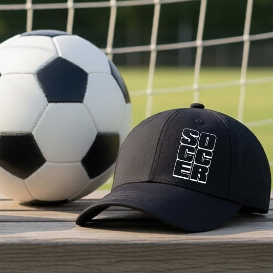 A black baseball cap with 'SOCCER' printed vertically in white on the front, resting on a wooden surface next to a classic black-and-white soccer ball with a blurred soccer net and grassy field in the background.