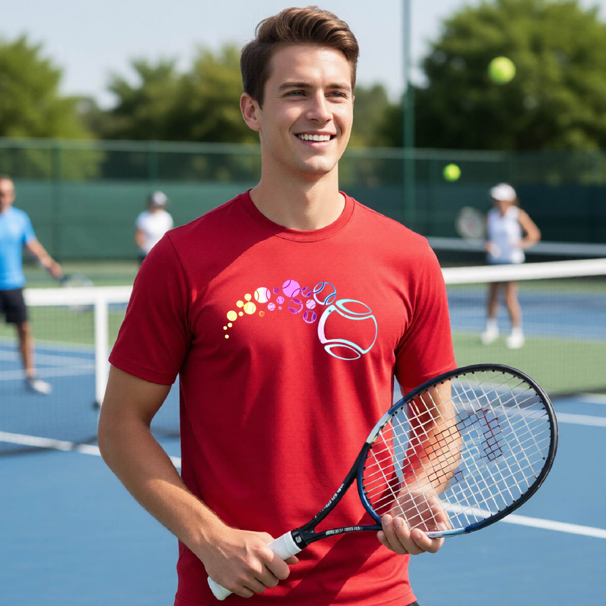 A man on a tennis court wearing a red short-sleeve t-shirt with a graphic of pastel gradient colored tennis balls arcing in flight and gradually getting larger.
