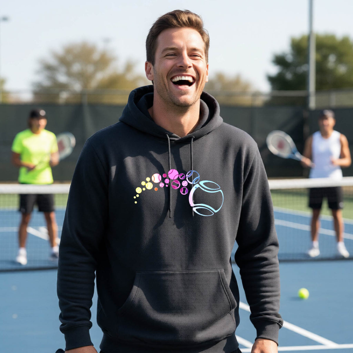 A man on a tennis court wearing a black hoodie pullover with a graphic of pastel gradient colored tennis balls arcing in flight and gradually getting larger.