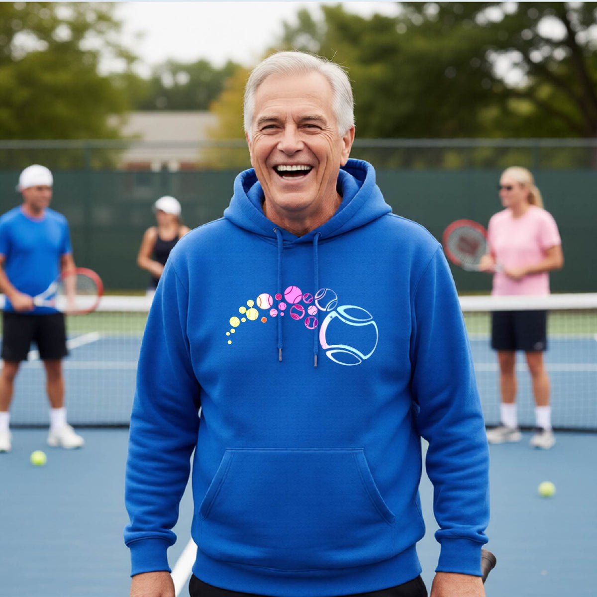 A man on a tennis court wearing a royal blue hoodie pullover with a graphic of pastel gradient colored tennis balls arcing in flight and gradually getting larger.