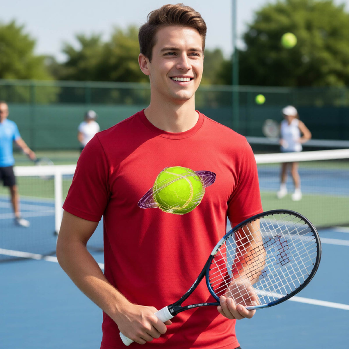 A man on a tennis court wearing a red short-sleeve t-shirt with a graphic of a tennis ball in orbit, holding a tennis racket. Other players are visible in the background on the court.