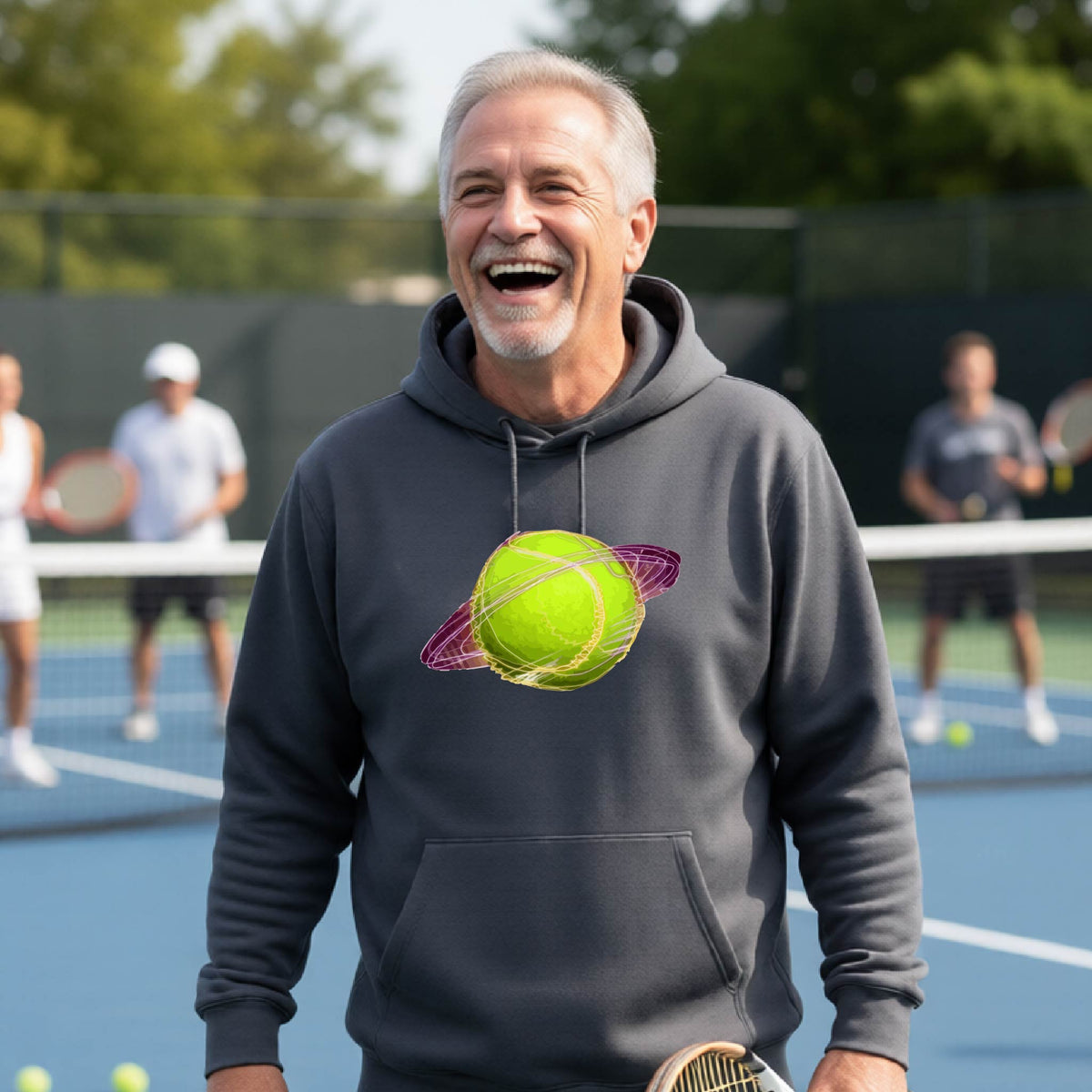 A man on a tennis court wearing a dark gray hoodie pullover with a graphic of a tennis ball in orbit, holding a tennis racket. Other players are visible in the background on the court.