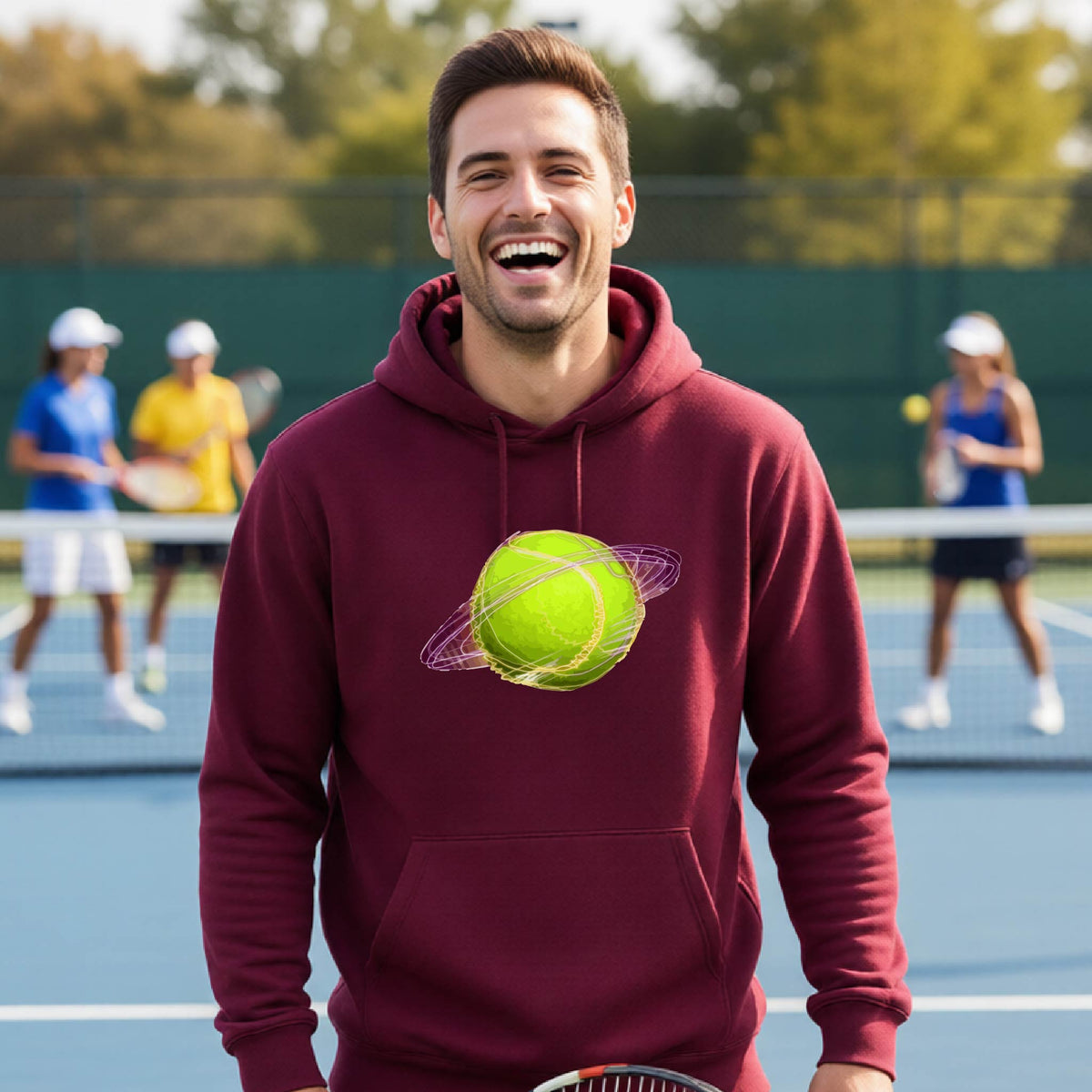 A man on a tennis court wearing a maroon hoodie pullover with a graphic of a tennis ball in orbit, holding a tennis racket. Other players are visible in the background on the court.