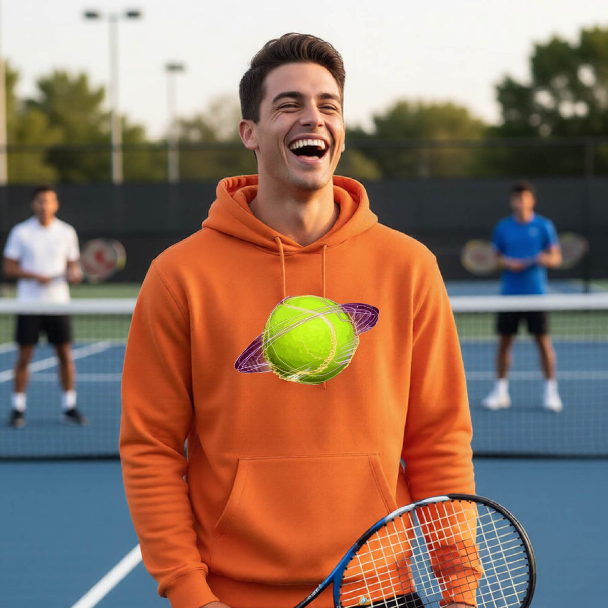 A man on a tennis court wearing an orange hoodie pullover with a graphic of a tennis ball in orbit, holding a tennis racket. Other players are visible in the background on the court.