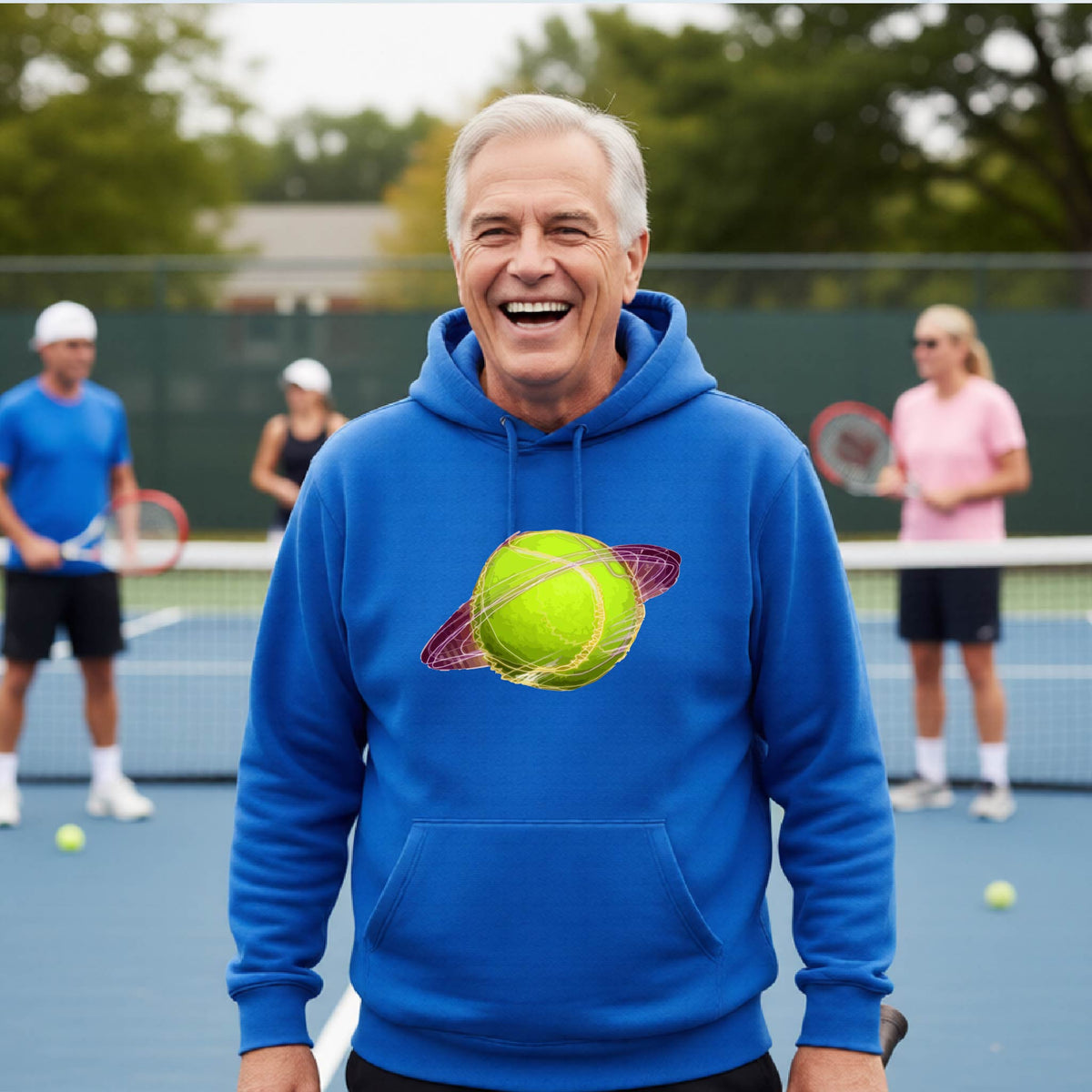 A man on a tennis court wearing a royal blue hoodie pullover with a graphic of a tennis ball in orbit, holding a tennis racket. Other players are visible in the background on the court.