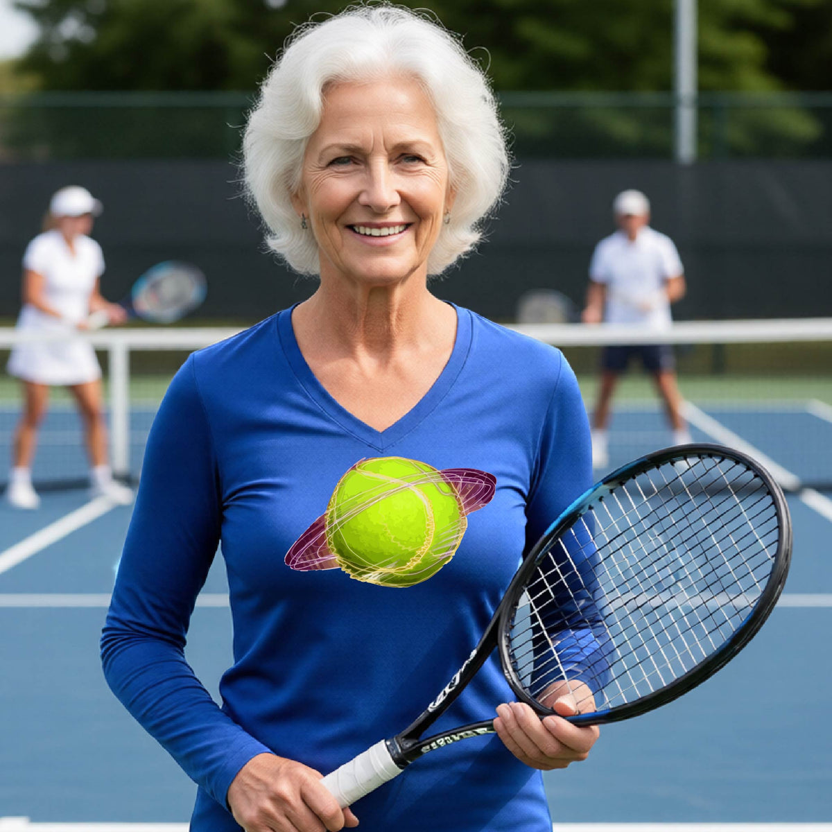 A woman on a tennis court wearing a royal blue long-sleeve t-shirt with a graphic of a tennis ball in orbit, holding a tennis racket. Other players are visible in the background on the court.