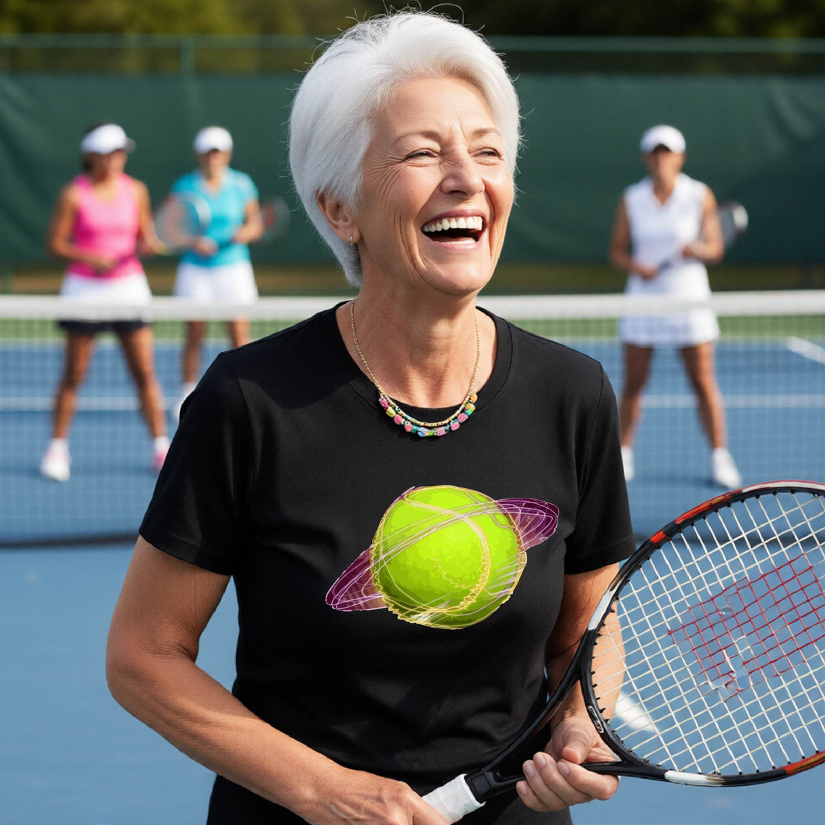 A woman on a tennis court wearing a black short-sleeve t-shirt with a graphic of a tennis ball in orbit, holding a tennis racket. Other players are visible in the background on the court.