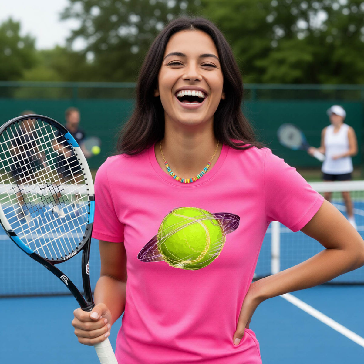 A woman on a tennis court wearing a pink short-sleeve t-shirt with a graphic of a tennis ball in orbit, holding a tennis racket. Other players are visible in the background on the court.