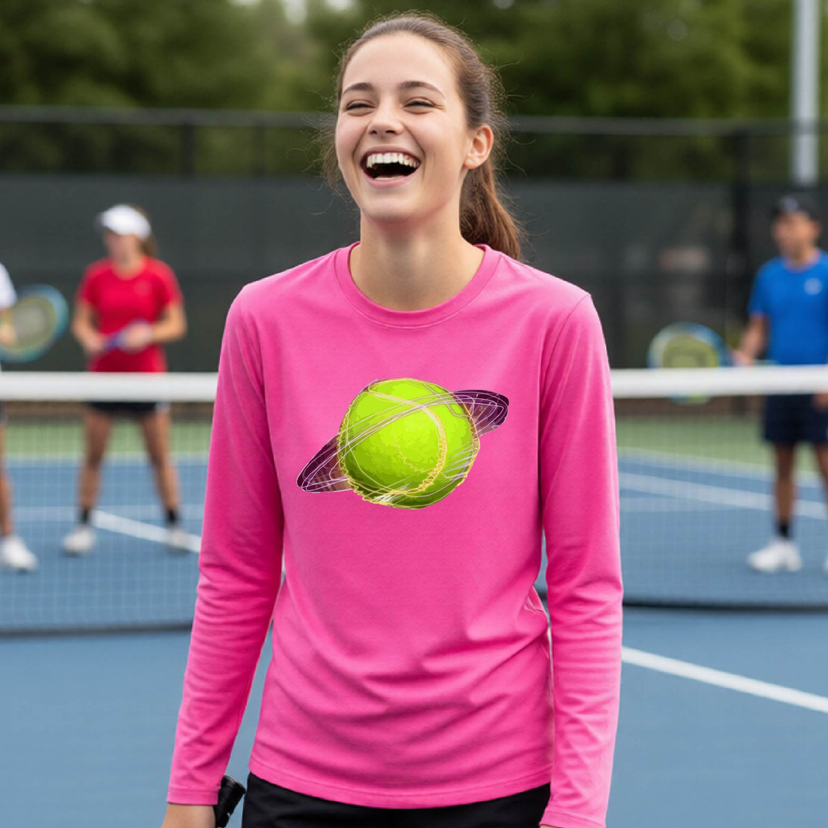 A young person on a tennis court wearing a pink long-sleeve t-shirt with a graphic of a tennis ball in orbit, holding a tennis racket. Other players are visible in the background on the court.