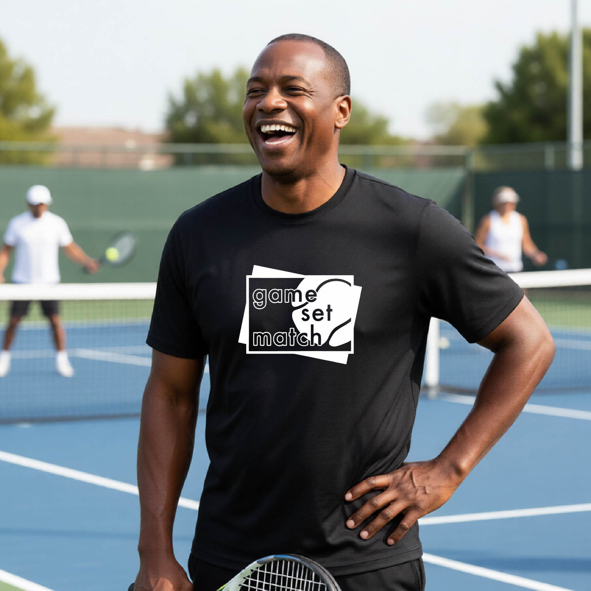 A man on a blue tennis court holds a tennis racket and wears a black short-sleeve T-shirt with a white graphic of two overlapping white and gray squares with the cut out words of ‘GAME SET MATCH” and a tennis ball, printed on the front of the shirt. 