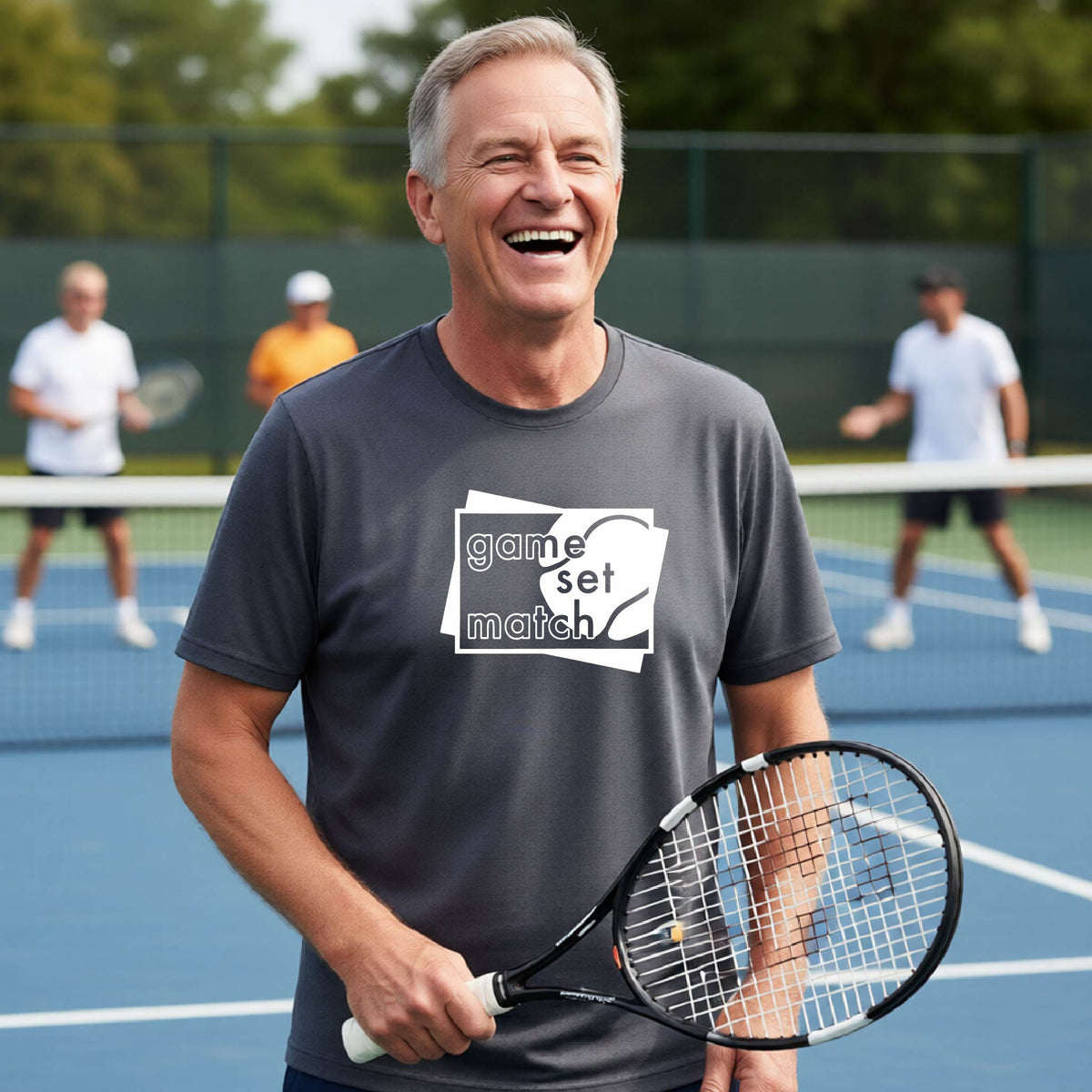 A man on a blue tennis court holds a tennis racket and wears a dark gray short-sleeve T-shirt with a white graphic of two overlapping white and gray squares with the cut out words of ‘GAME SET MATCH” and a tennis ball, printed on the front of the shirt. 