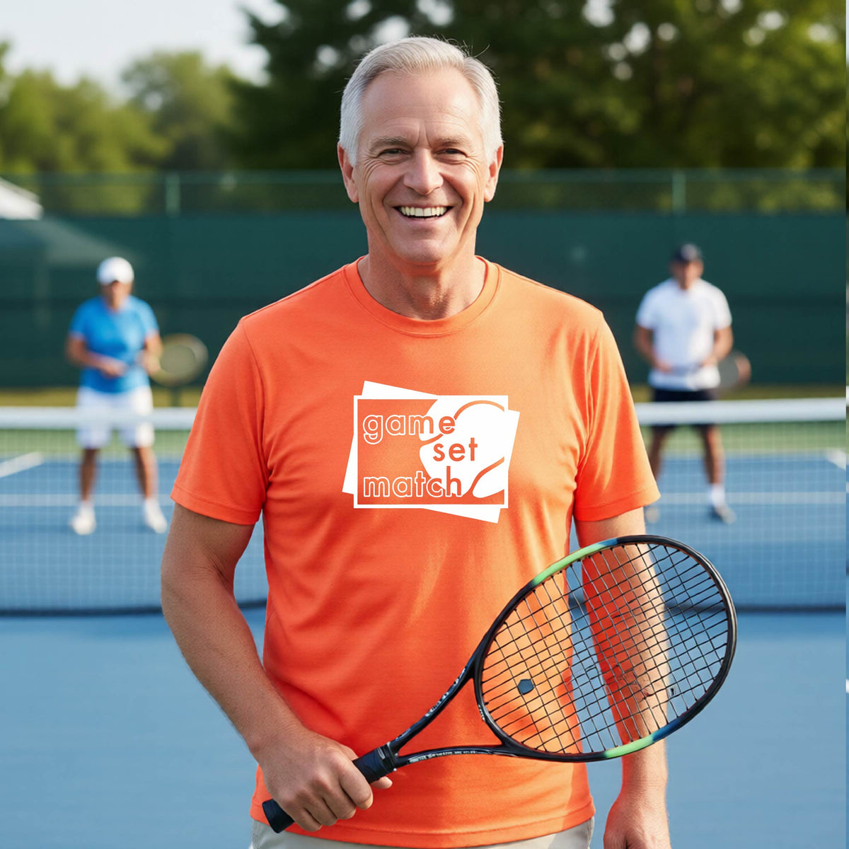 A man on a blue tennis court holds a tennis racket and wears an orange short-sleeve T-shirt with a white graphic of two overlapping white and gray squares with the cut out words of ‘GAME SET MATCH” and a tennis ball, printed on the front of the shirt. 