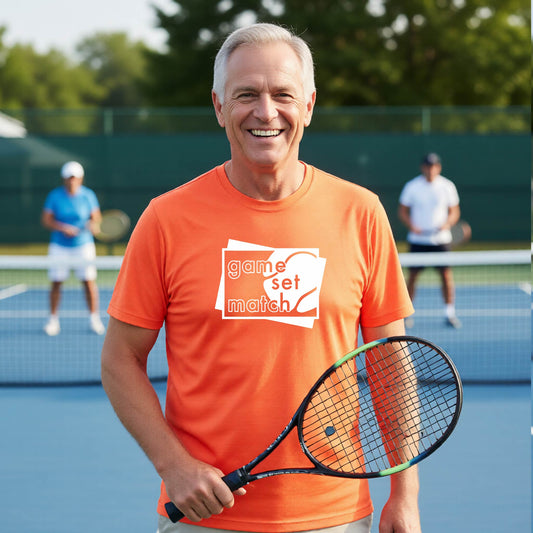 A man on a blue tennis court holds a tennis racket and wears an orange short-sleeve T-shirt with a white graphic of two overlapping white and gray squares with the cut out words of ‘GAME SET MATCH” and a tennis ball, printed on the front of the shirt. 