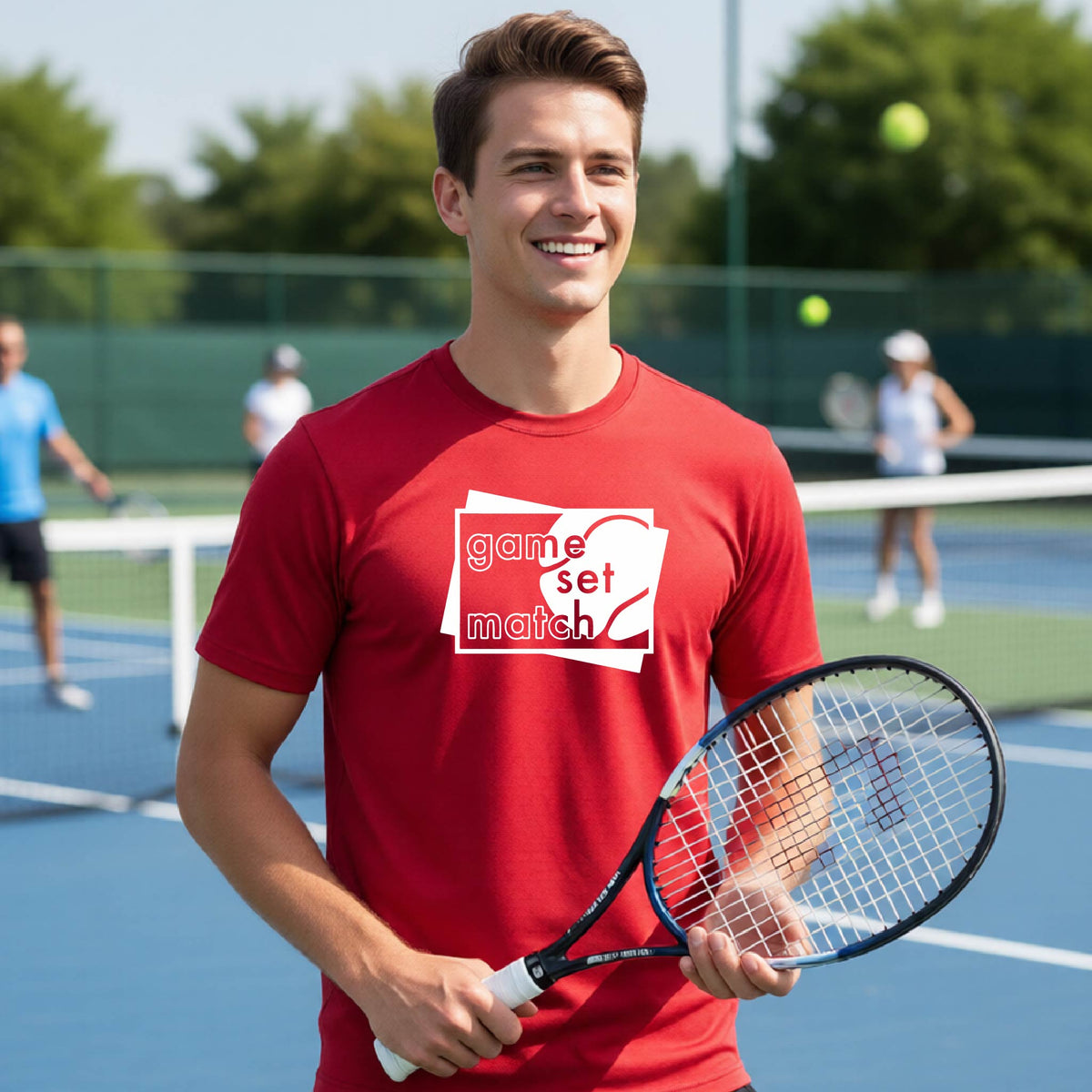A man on a blue tennis court holds a tennis racket and wears a red short-sleeve T-shirt with a white graphic of two overlapping white and gray squares with the cut out words of ‘GAME SET MATCH” and a tennis ball, printed on the front of the shirt. 