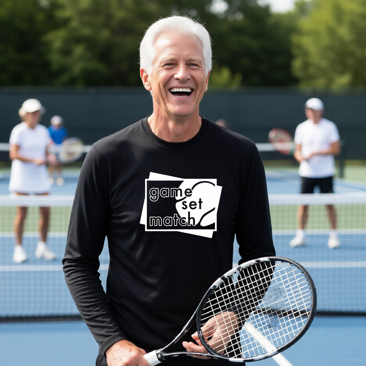 A man on a blue tennis court holds a tennis racket and wears a black long-sleeve T-shirt with a white graphic of two overlapping white and gray squares with the cut out words of ‘GAME SET MATCH” and a tennis ball, printed on the front of the shirt. 